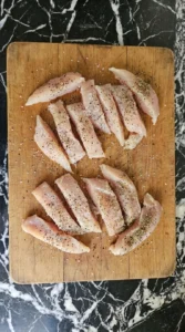 Uneven chicken strips seasoned for chicken alfredo on a cutting board.