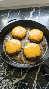 Melted sharp cheddar cheese draping over cooked burger patties in a skillet