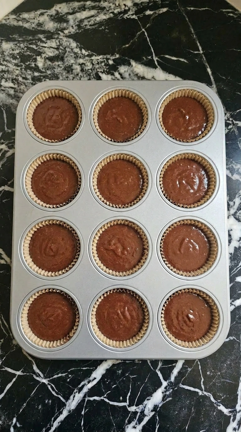 Chocolate cupcake batter divided evenly into pleated paper liners inside an aluminum muffin tin