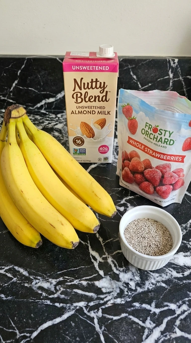 Raw ingredients for fruit smoothies scattered across a counter including bananas, frozen strawberries, and almond milk.