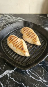 Two chicken breasts cooking in a cast iron grill pan with distinct dark char marks visible on the surface