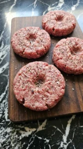 Seasoning raw burger patties with salt and pepper on a cutting board