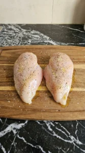 Two raw boneless skinless chicken breasts being seasoned with salt pepper and garlic powder on a wooden cutting board