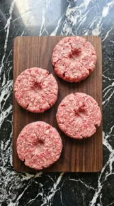 Four raw ground beef burger patties with center dimples on a wooden board