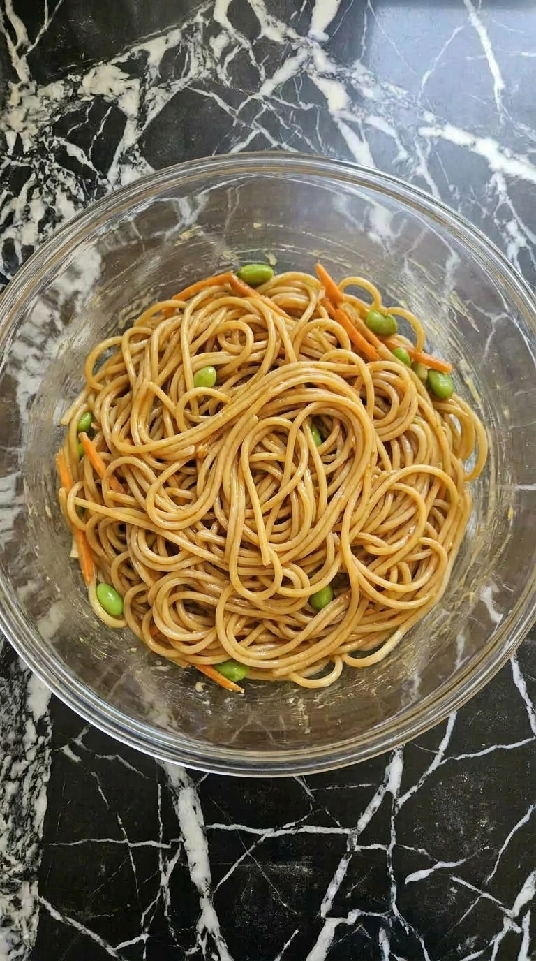 Spaghetti noodles being tossed with peanut dressing in a glass mixing bowl
