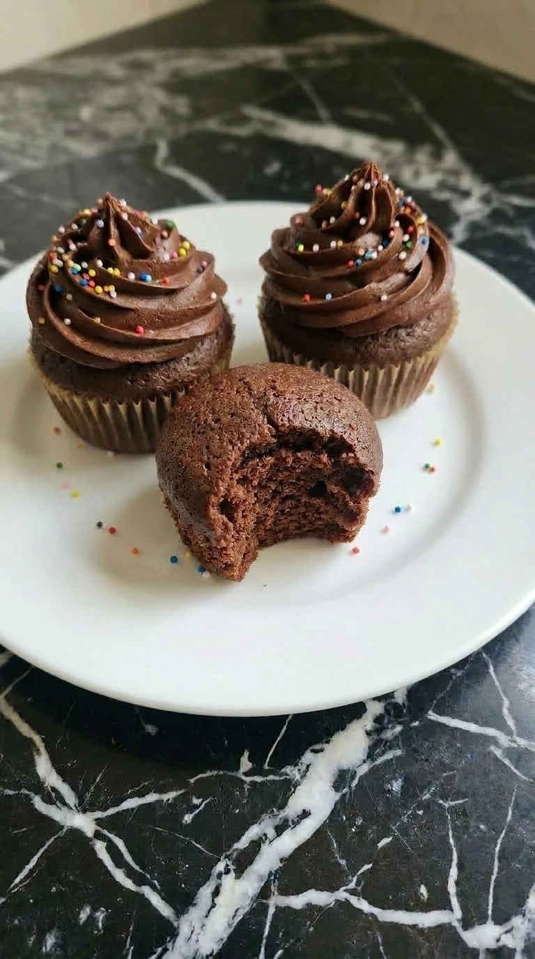 Three rich dark chocolate cupcakes with deep brown buttercream frosting and rainbow sprinkles on a white plate