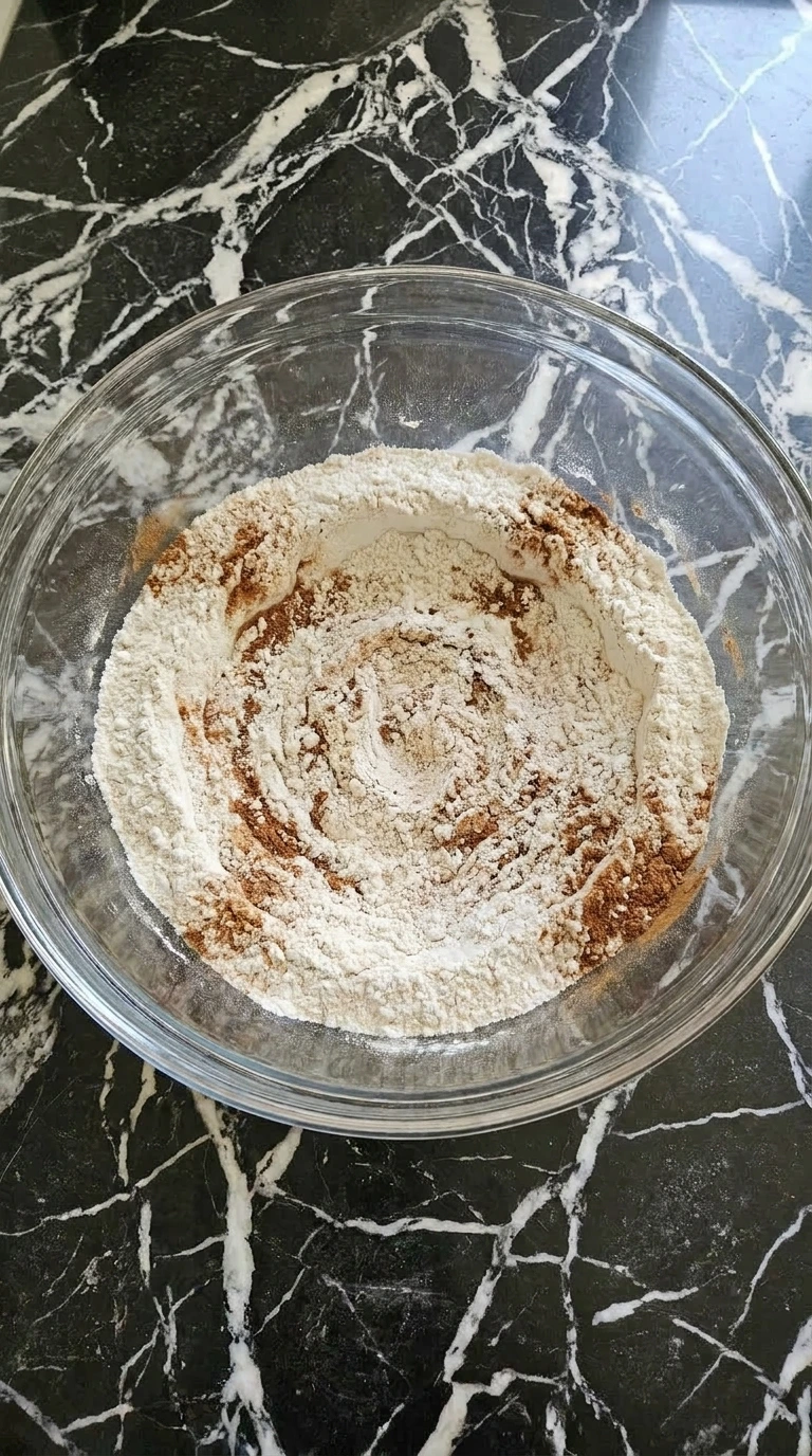 Flour and autumn baking spices mixed together inside a large clear glass mixing bowl
