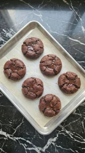 Six freshly baked double chocolate chip cookies sitting hot on a parchment lined sheet pan