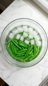 Vibrant green french green beans cooling inside a glass bowl filled with ice water.