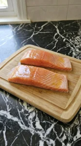 Two raw pink salmon fillets being prepped with olive oil on a wooden board