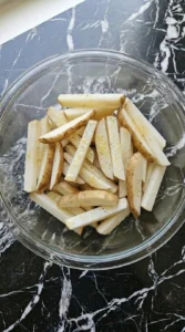Oil-coated and seasoned raw potato sticks inside a clear glass mixing bowl.