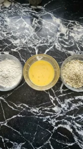 Three shallow glass bowls holding flour, whisked egg, and seasoned panko breadcrumbs side by side.