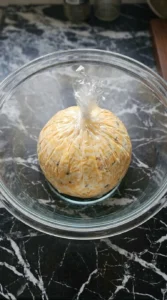 A round cheese ball wrapped tightly in clear plastic wrap sitting inside a glass bowl.