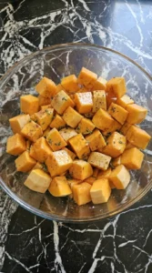 Raw butternut squash cubes coated with maple glaze and seasonings in a glass bowl.