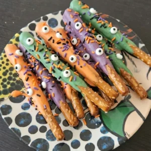 Halloween Party Food Pretzels with colorful chocolate coatings, candy eyes and orange black and purple jimmies on a white platter